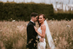 Bride and Groom in long grass as bride stares to camera.